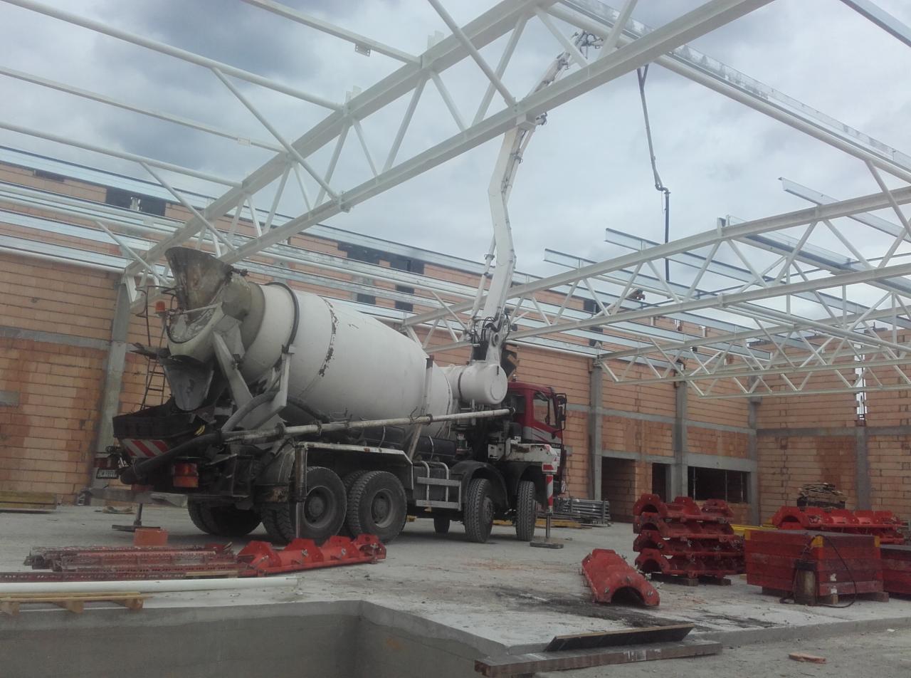 Ciężarówka z betonomieszarką i wysięgnikiem pompy betonu na placu budowy, widoczne elementy konstrukcji stalowej dachu i ceglane ściany w tle.