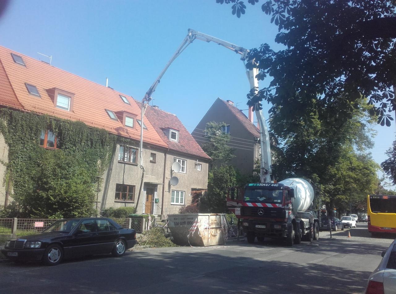 Ciężarówka z pompą do betonu Coliner 440 na tle dwupiętrowych domów z czerwonymi dachami, jeden z domów porośnięty bluszczem, obok czarny Mercedes i kontener na gruz.