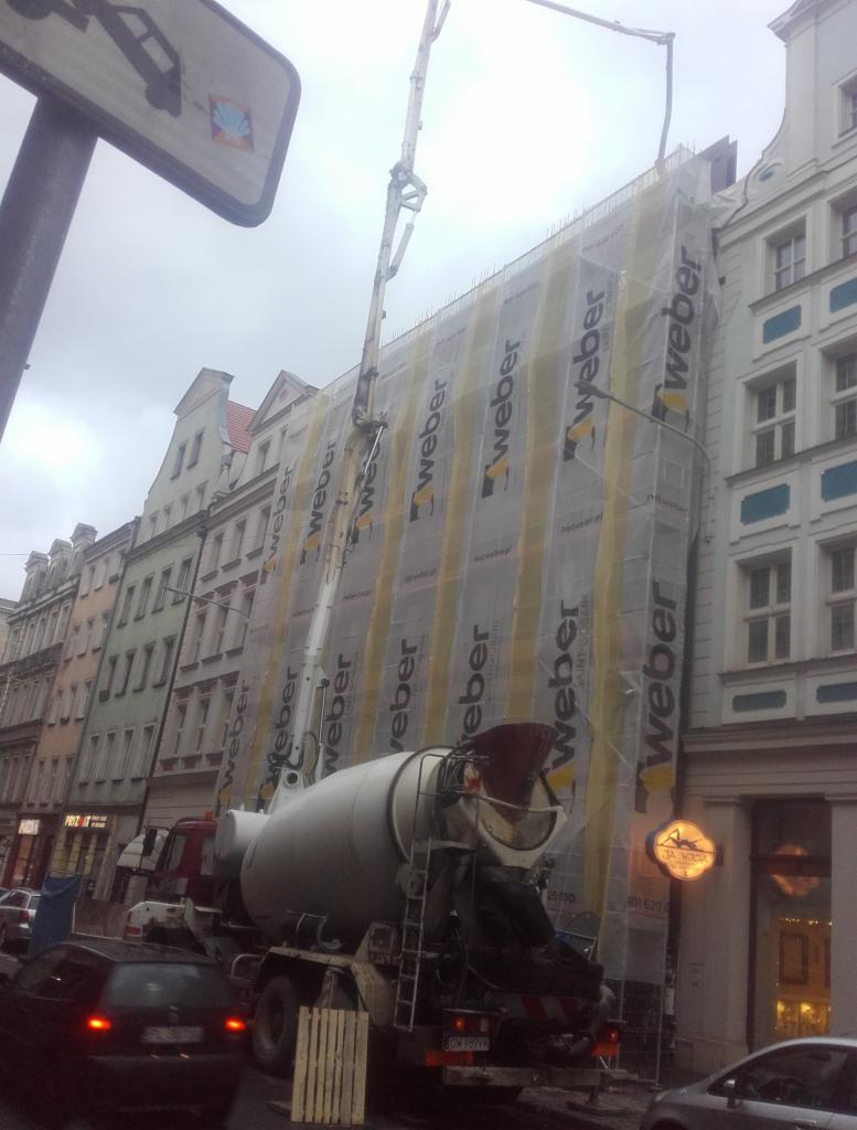 Betonomieszarka z wysięgnikiem pompuje beton na budynek w rusztowaniach, zabezpieczony siatką z logo Weber, na tle kamienic w pochmurny dzień.