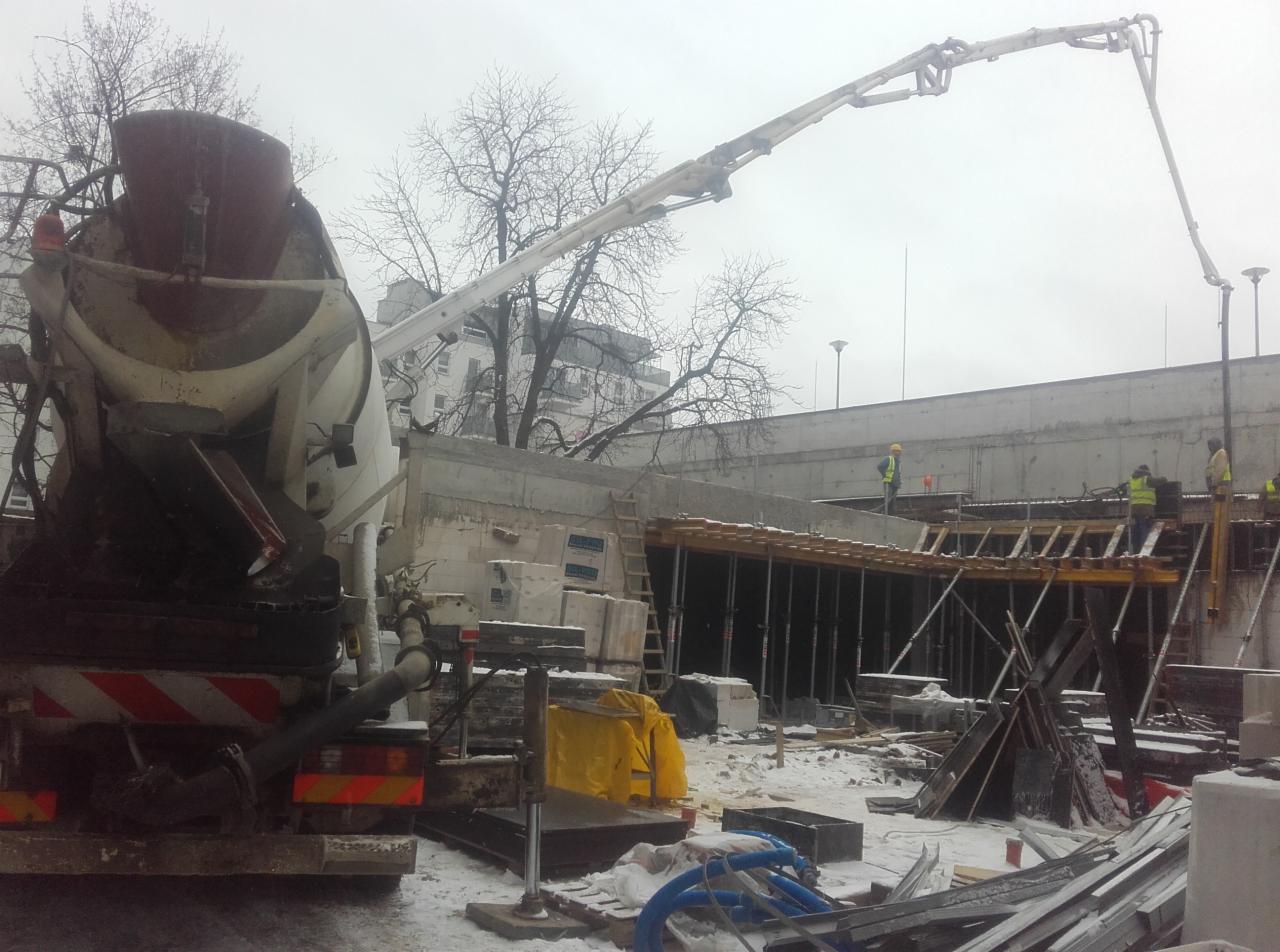 Betonomieszarka z pompą podaje beton na budowę w zimowy dzień, widoczni pracownicy w żółtych kamizelkach, szalunki przygotowane do wylewania betonu.