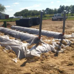 Montaż drenażu rozsączającego przydomowej oczyszczalni ścieków, rury drenażowe owinięte geowłókniną, wykop z piaskiem, w tle zbiorniki oczyszczalni i koparka.