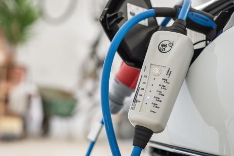 Biała ładowarka do samochodu elektrycznego podłączona do gniazda, z niebieskim kablem i diodami LED wskazującymi poziom naładowania.