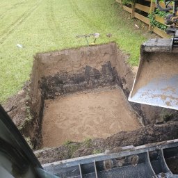 Max-Bud - Wykopany dół w ziemi z widoczną warstwą piasku na dnie, obok stoi koparka. W tle trawa i drewniany płot.