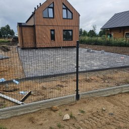 Max-Bud - Nowoczesny dom z drewnianą elewacją, otoczony szarą kostką brukową i czarnym ogrodzeniem panelowym z betonową podmurówką. Widoczne przygotowania do dalszych prac.