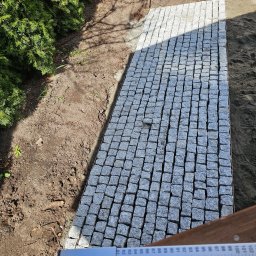 Max-Bud - Fragment ścieżki z kostki granitowej, widok z góry, zarysowany kształt, obrzeża z metalu, częściowo ułożona, obok ziemia i trawa, w tle ogrodzenie.