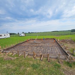 Max-Bud - Zbrojenie fundamentu pod budynek, widoczne szalunki i siatka zbrojeniowa na folii, w tle łąka i dwa białe kampery. Prace przygotowawcze do wylania betonu.