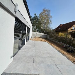 Max-Bud - Nowoczesny taras z szarych płyt, obrzeża z kostki. Widok na dom z dużymi oknami i ogród w tle. Częściowo ukończone prace, widoczne narzędzia i materiały.