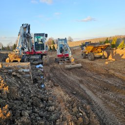 Max-Bud - Dwa biało-czerwone mini-koparki i żółta wywrotka na placu budowy, świeżo wykopana ziemia, ślady opon, błękitne niebo z chmurami w tle, wczesny etap prac ziemnych.