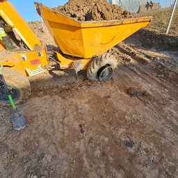 Max-Bud - Żółta wywrotka z ładunkiem ziemi na placu budowy, widoczna łopata i fragment ogrodzenia. Ślady opon na ziemi sugerują prace ziemne.