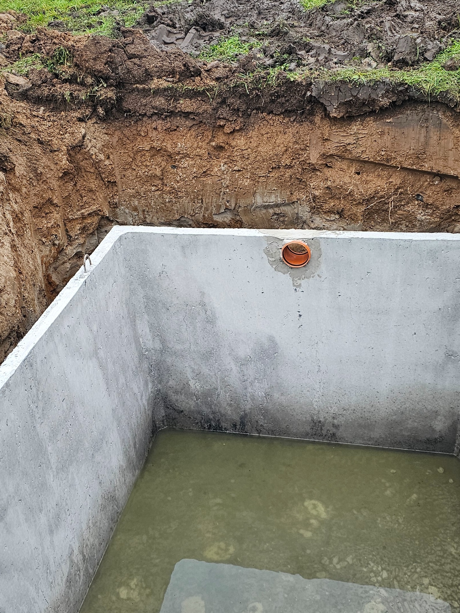 Betonowy zbiornik z otworem na rurę, częściowo wypełniony wodą, umieszczony w wykopie ziemnym. Widoczna struktura gruntu i trawa na górze.