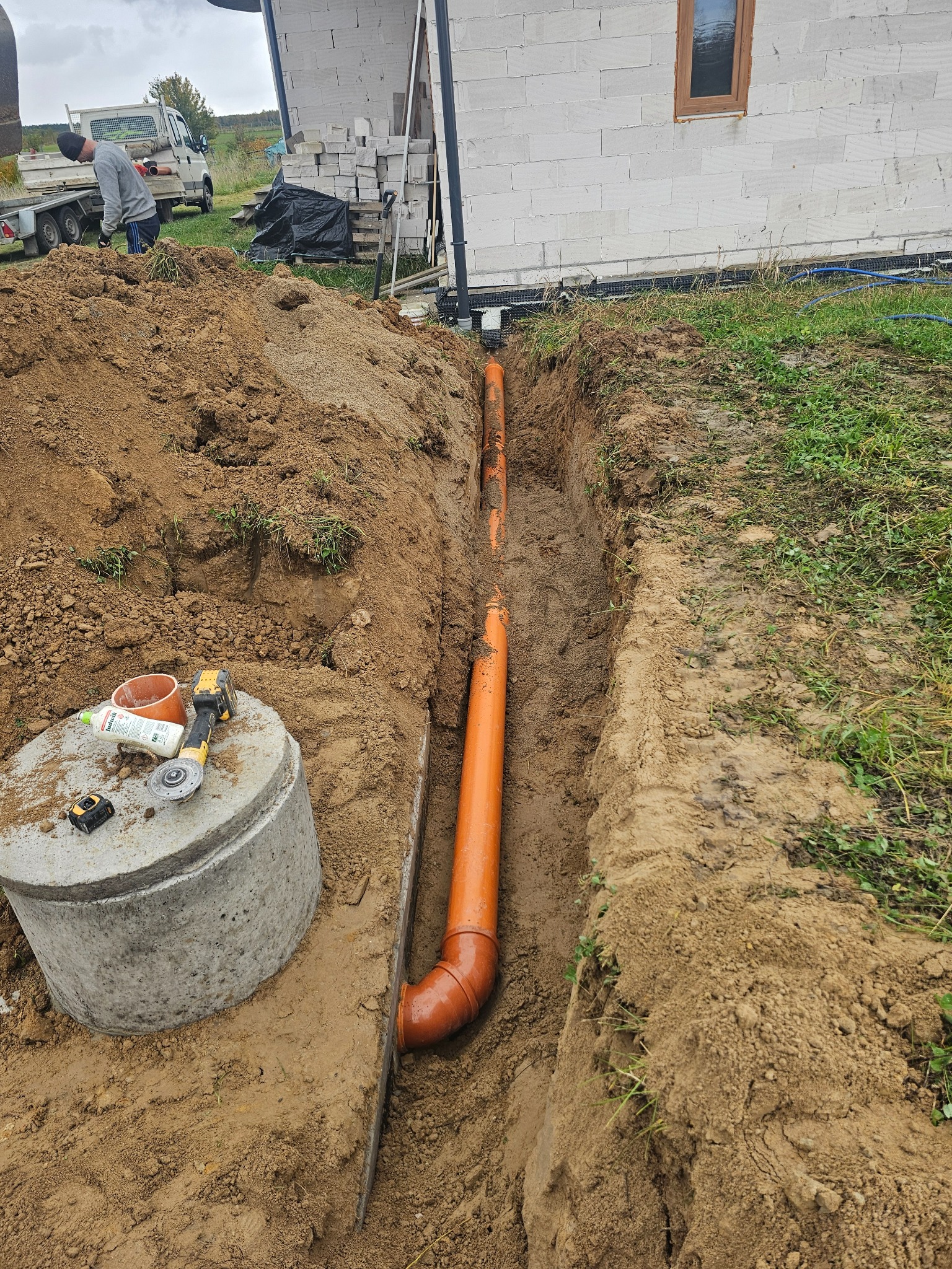 Pomarańczowe rury ułożone w wykopie, betonowy krąg studni z narzędziami, w tle budynek w trakcie budowy. Widok z góry na instalację kanalizacyjną.