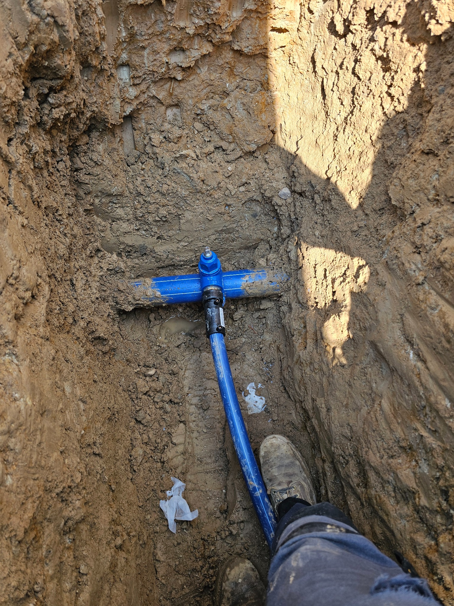 Niebieskie rury wodociągowe w wykopie z ziemi, widoczne z góry, z fragmentem buta pracownika. Instalacja wodna w trakcie montażu.
