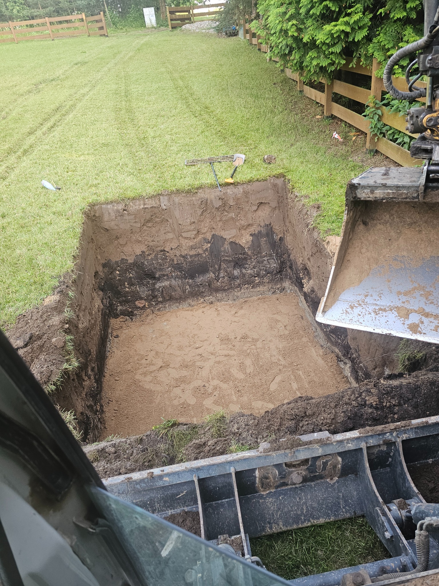 Wykopany dół w ziemi z widoczną warstwą piasku na dnie, obok stoi koparka. W tle trawa i drewniany płot.