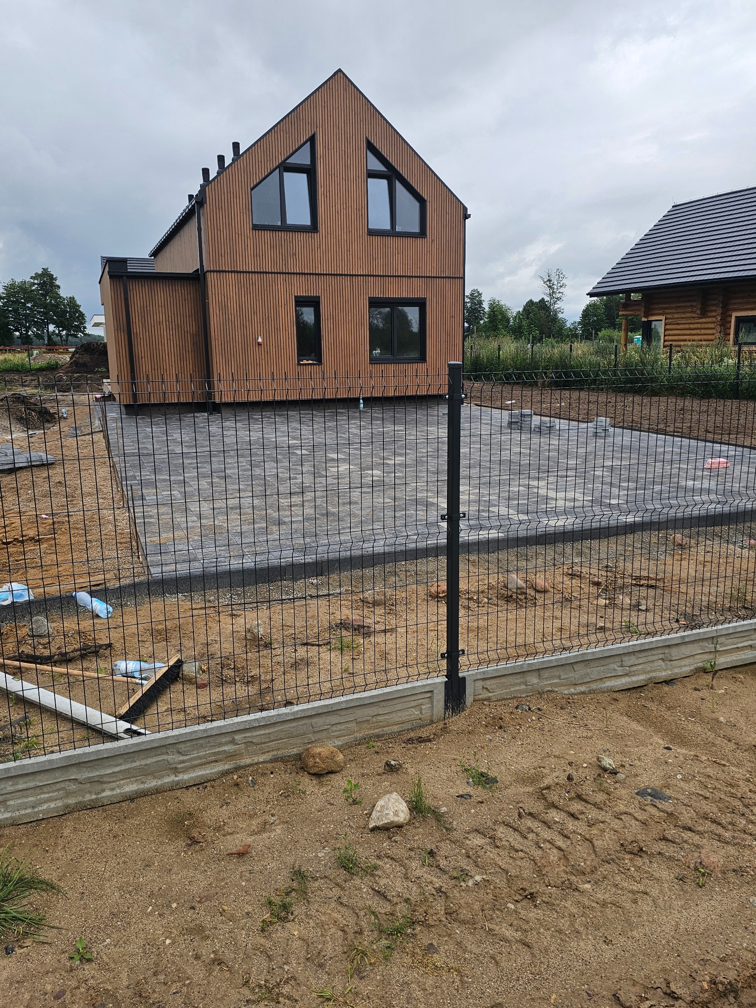 Nowoczesny dom z drewnianą elewacją, otoczony szarą kostką brukową i czarnym ogrodzeniem panelowym z betonową podmurówką. Widoczne przygotowania do dalszych prac.