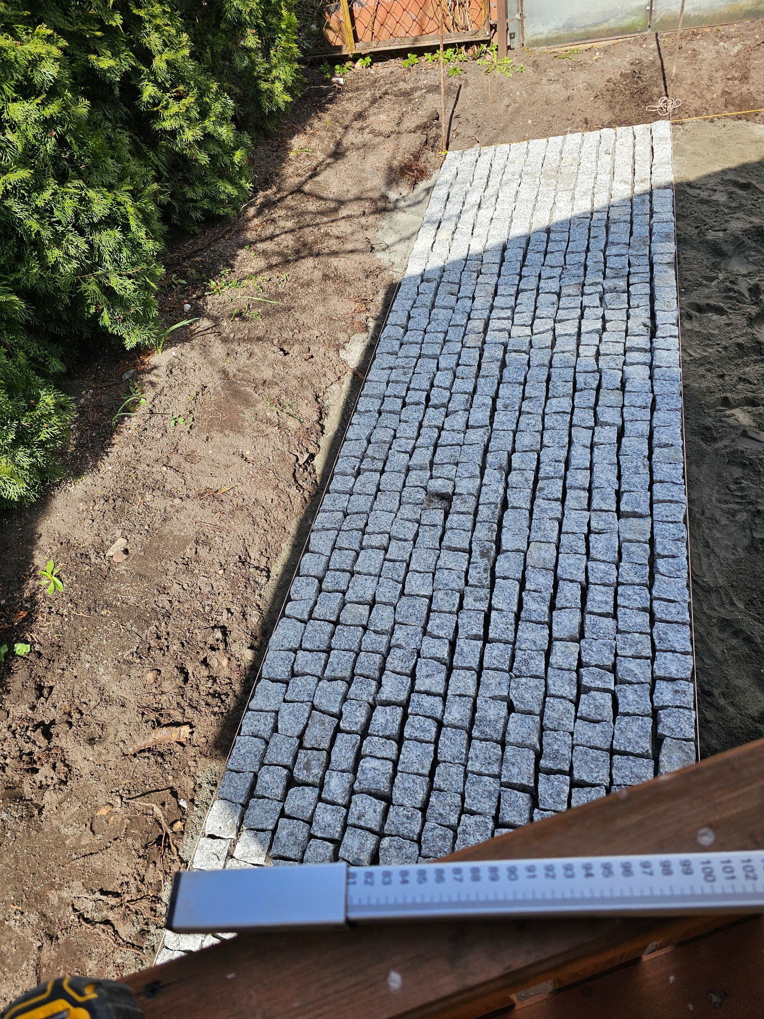 Fragment ścieżki z kostki granitowej, widok z góry, zarysowany kształt, obrzeża z metalu, częściowo ułożona, obok ziemia i trawa, w tle ogrodzenie.