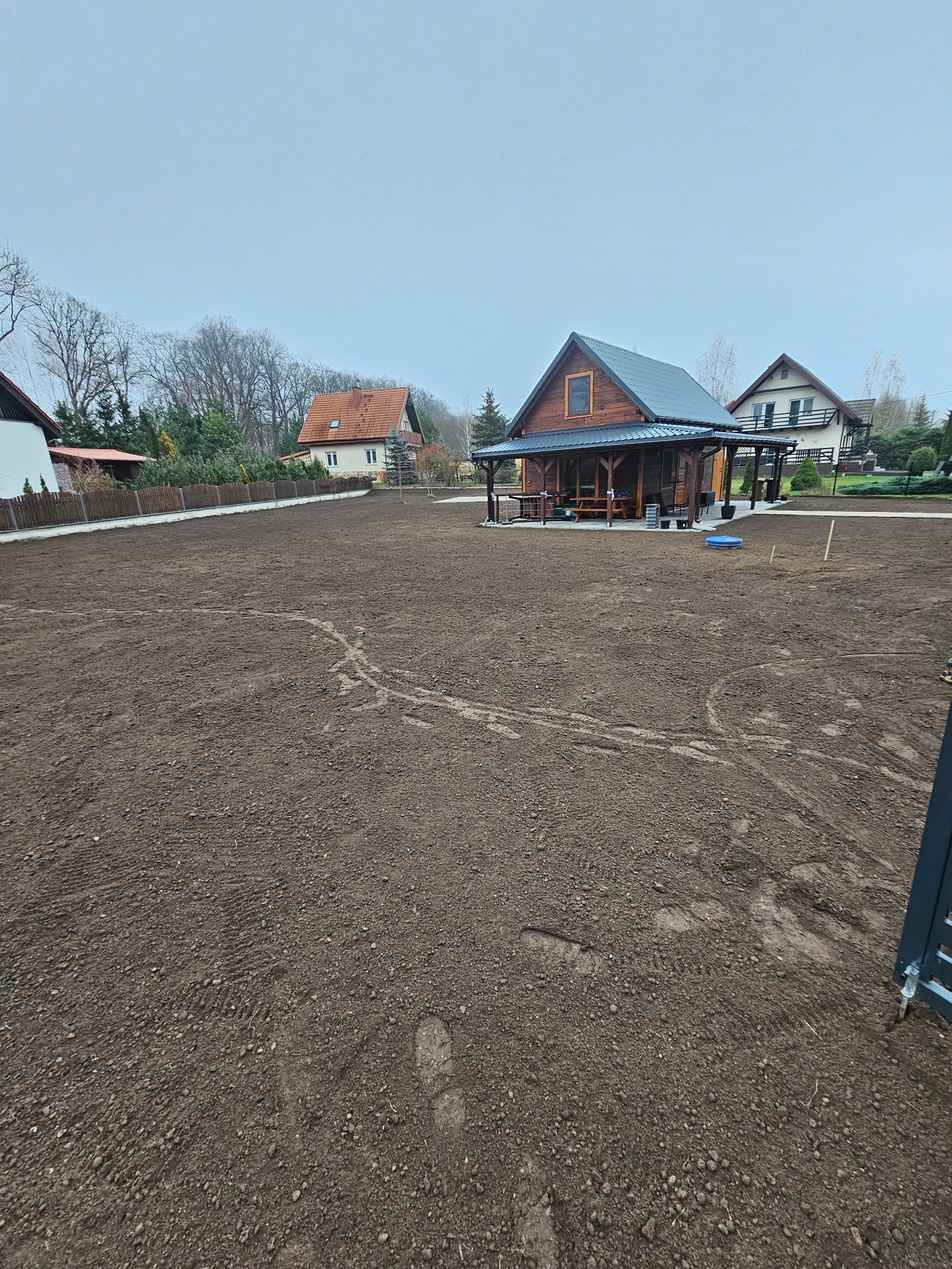 Przygotowany teren pod trawnik z widoczną drewnianą altaną i domami w tle. Ślady opon na ziemi sugerują niedawne prace ziemne.