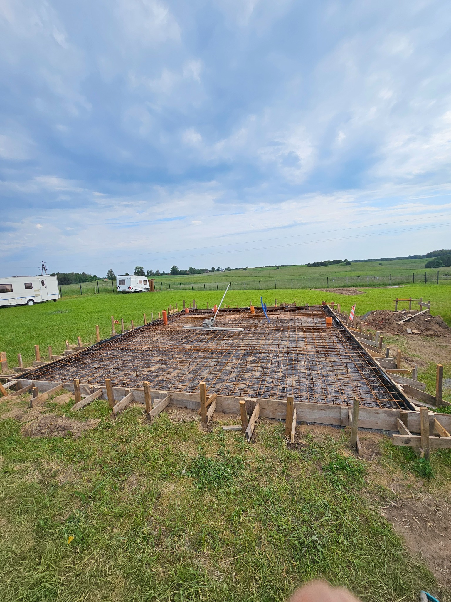 Zbrojenie fundamentu pod budynek, widoczne szalunki i siatka zbrojeniowa na folii, w tle łąka i dwa białe kampery. Prace przygotowawcze do wylania betonu.