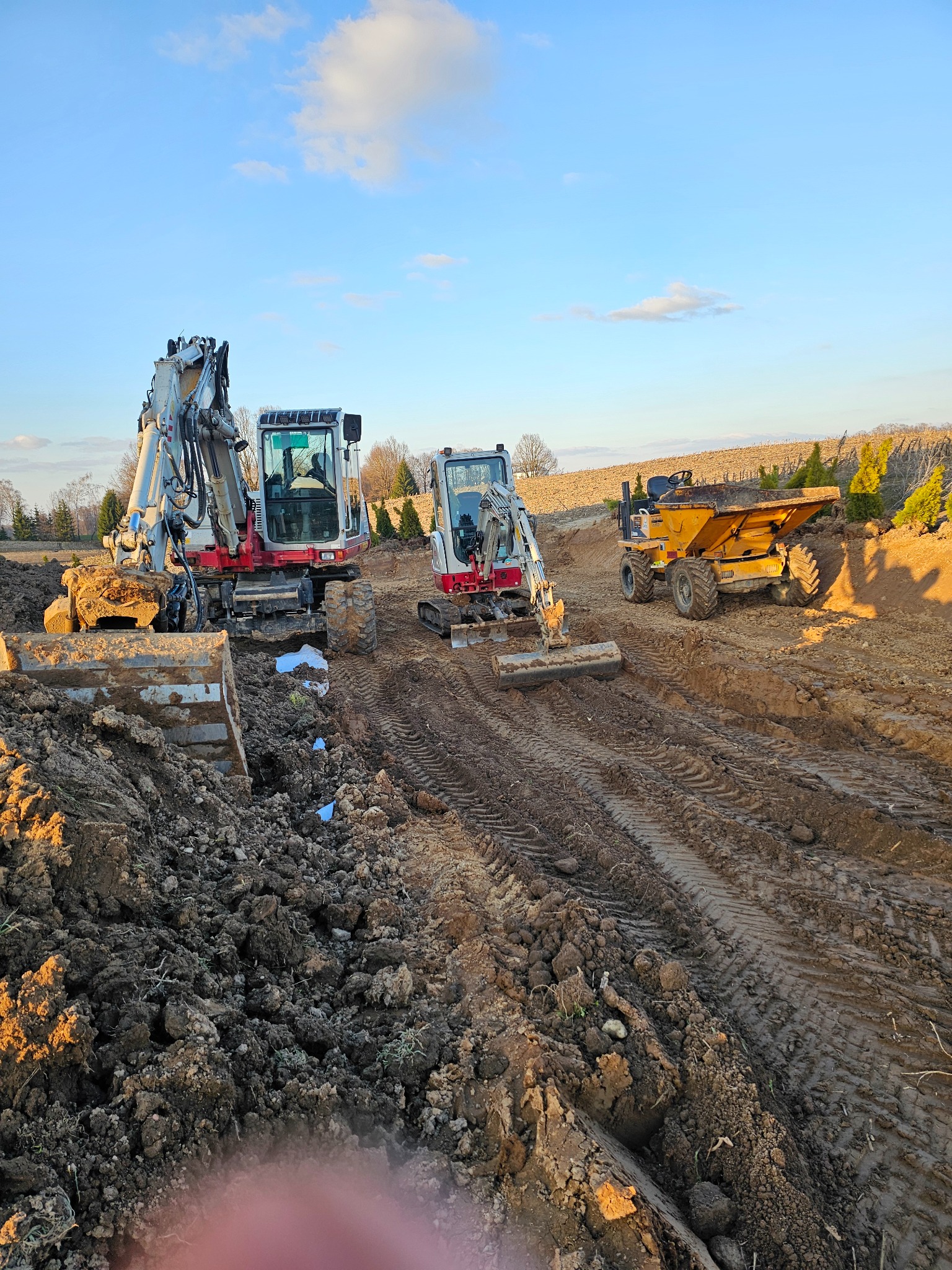 Dwa biało-czerwone mini-koparki i żółta wywrotka na placu budowy, świeżo wykopana ziemia, ślady opon, błękitne niebo z chmurami w tle, wczesny etap prac ziemnych.