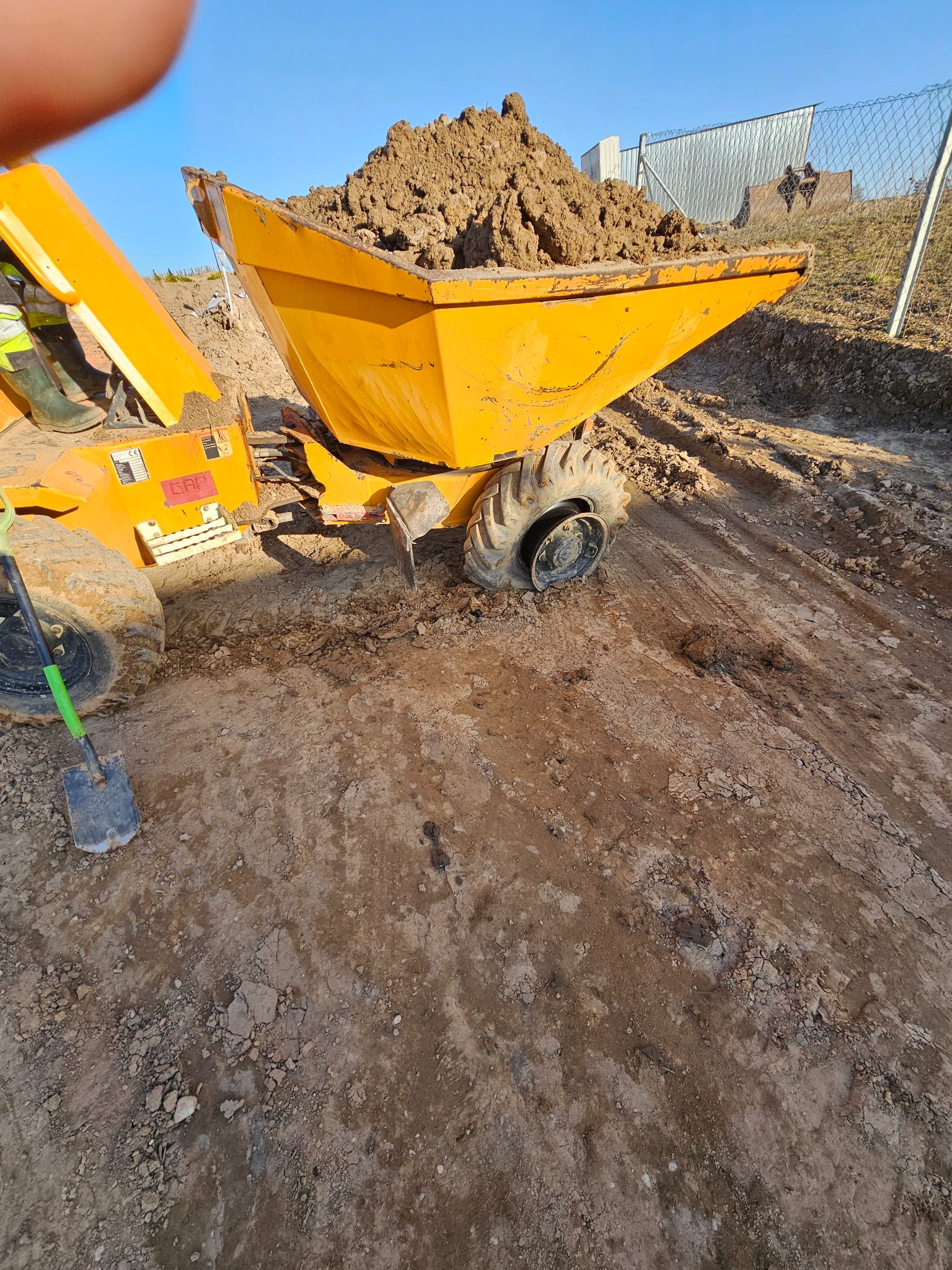Żółta wywrotka z ładunkiem ziemi na placu budowy, widoczna łopata i fragment ogrodzenia. Ślady opon na ziemi sugerują prace ziemne.