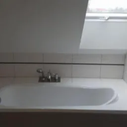 Biała wanna obudowana szarą płytką, bateria z prysznicem, białe płytki na ścianach z delikatnym wzorem fal, okno dachowe w skosie.