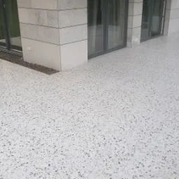 Jasnoszara, polerowana posadzka terrazzo na zewnątrz budynku z dużymi oknami, widoczne drobne kamyczki w strukturze, minimalistyczny design.