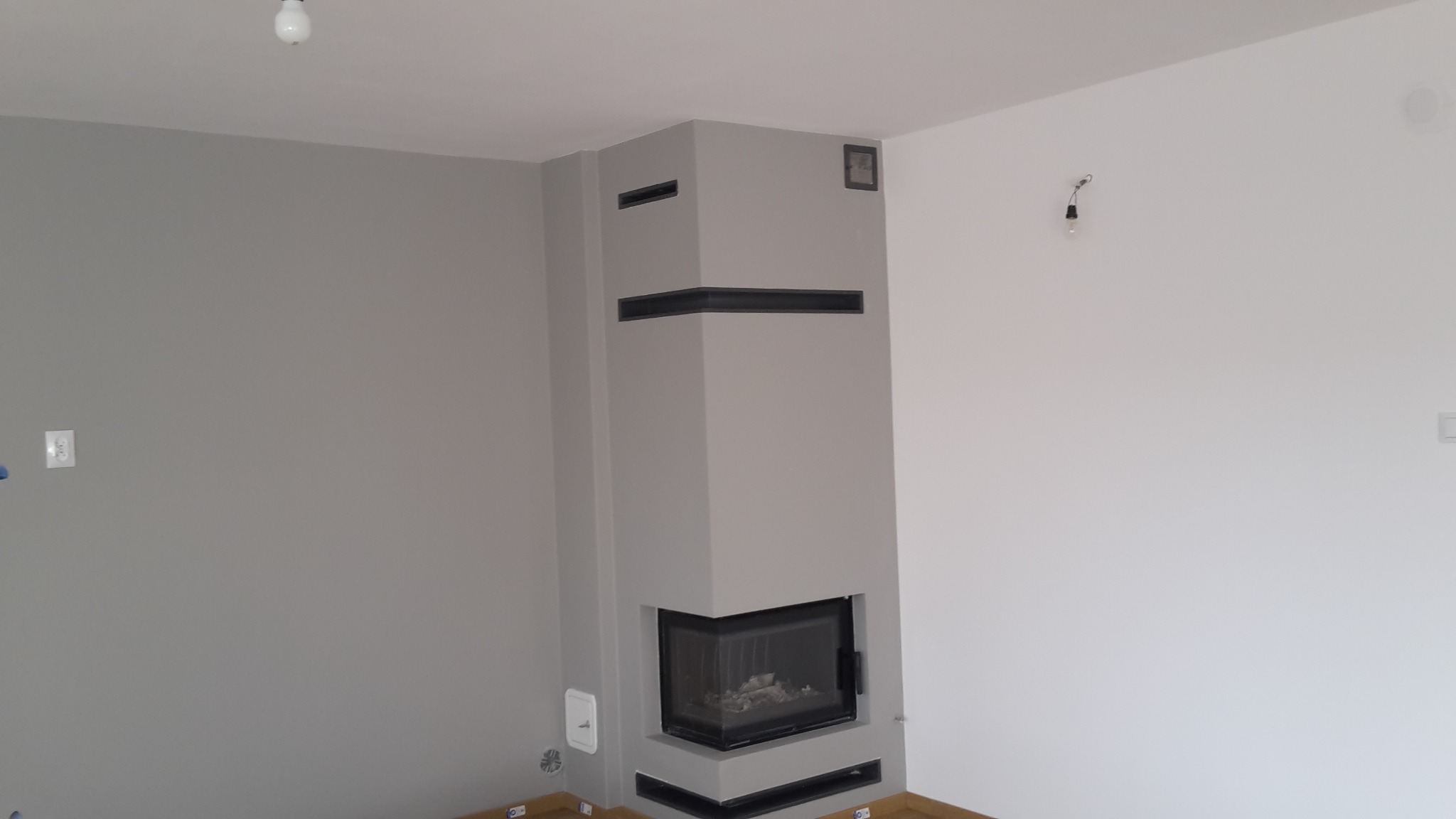 Szary kominek narożny z wkładem kominkowym, czarnymi elementami dekoracyjnymi i kratkami wentylacyjnymi, na tle szarej i białej ściany w surowym wnętrzu.