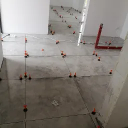 Szare płytki podłogowe w trakcie montażu, z użyciem poziomujących klipsów i klinów, widoczny poziomica, wnętrze w trakcie remontu.