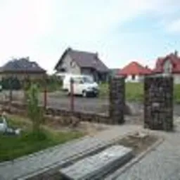 Fragment posesji z niedokończonym podjazdem z szarej kostki, widoczne palety z kostką, w tle dom jednorodzinny i zaparkowany biały bus.