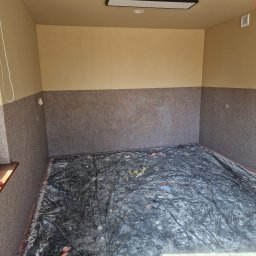 Marcin Nowakowski - Pokój po częściowym wykończeniu: dolna część ścian pokryta szarą strukturą, górna część beżowa. Podłoga zabezpieczona folią malarską. Widok na okno z parapetem.