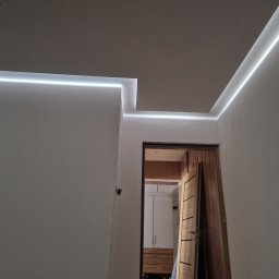 Marcin Nowakowski - Nowoczesne oświetlenie LED wzdłuż krawędzi sufitu i ścian, tworzące minimalistyczny design. Widok na otwarte drzwi do pomieszczenia z szafą.