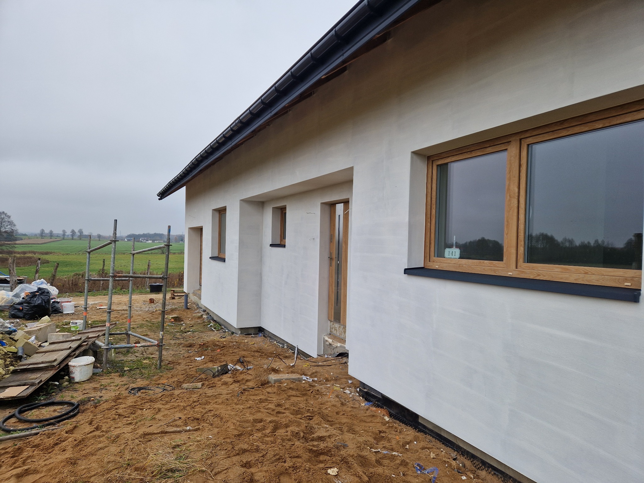 Elewacja domu jednorodzinnego w trakcie prac wykończeniowych, widoczne rusztowania, piasek i narzędzia. Świeżo otynkowana ściana z drewnianymi oknami.