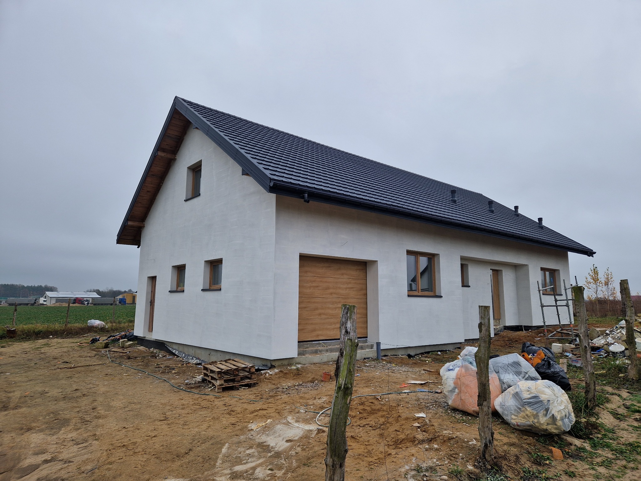 Nowoczesny dom jednorodzinny z ciemną dachówką i jasną elewacją, widoczny z zewnątrz, z elementami wykończeniowymi i workami z odpadami budowlanymi na pierwszym planie.
