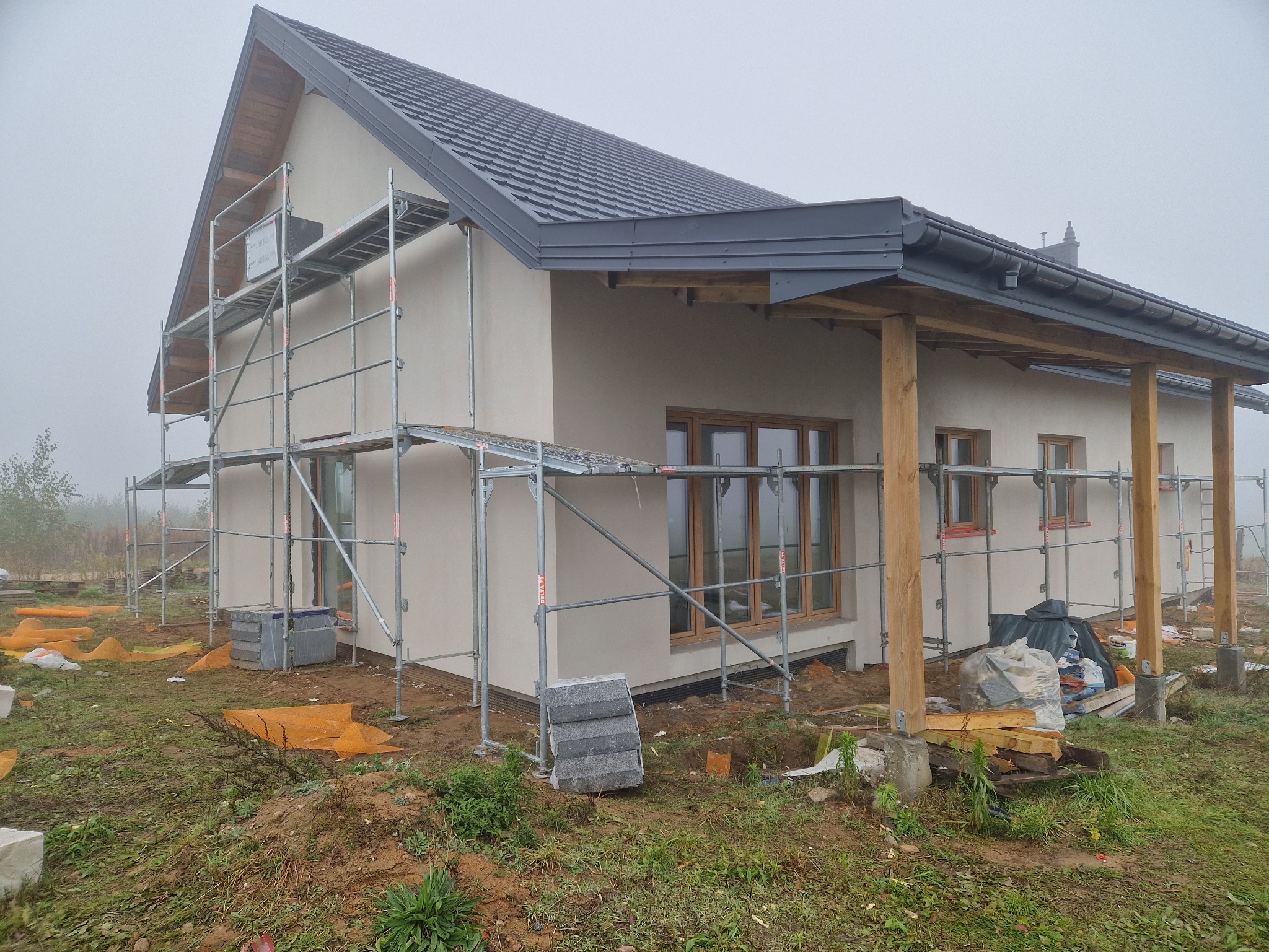 Dom jednorodzinny w trakcie prac wykończeniowych elewacji, z widocznym rusztowaniem i materiałami budowlanymi na posesji w pochmurny dzień.