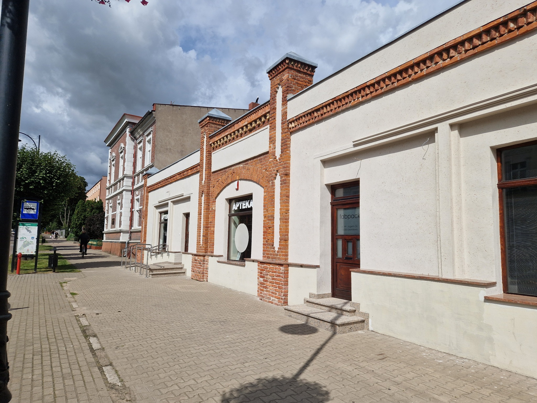Odnowiona fasada budynku z elementami ceglanymi i białym tynkiem, widok z chodnika w słoneczny dzień. Widoczny szyld apteki i wejście do lokalu.