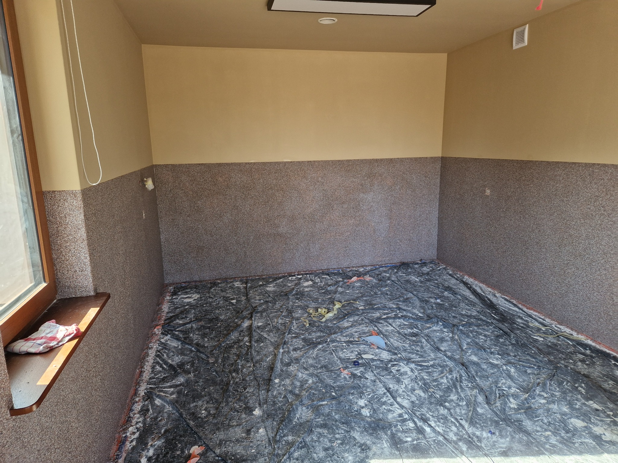 Pokój po częściowym wykończeniu: dolna część ścian pokryta szarą strukturą, górna część beżowa. Podłoga zabezpieczona folią malarską. Widok na okno z parapetem.