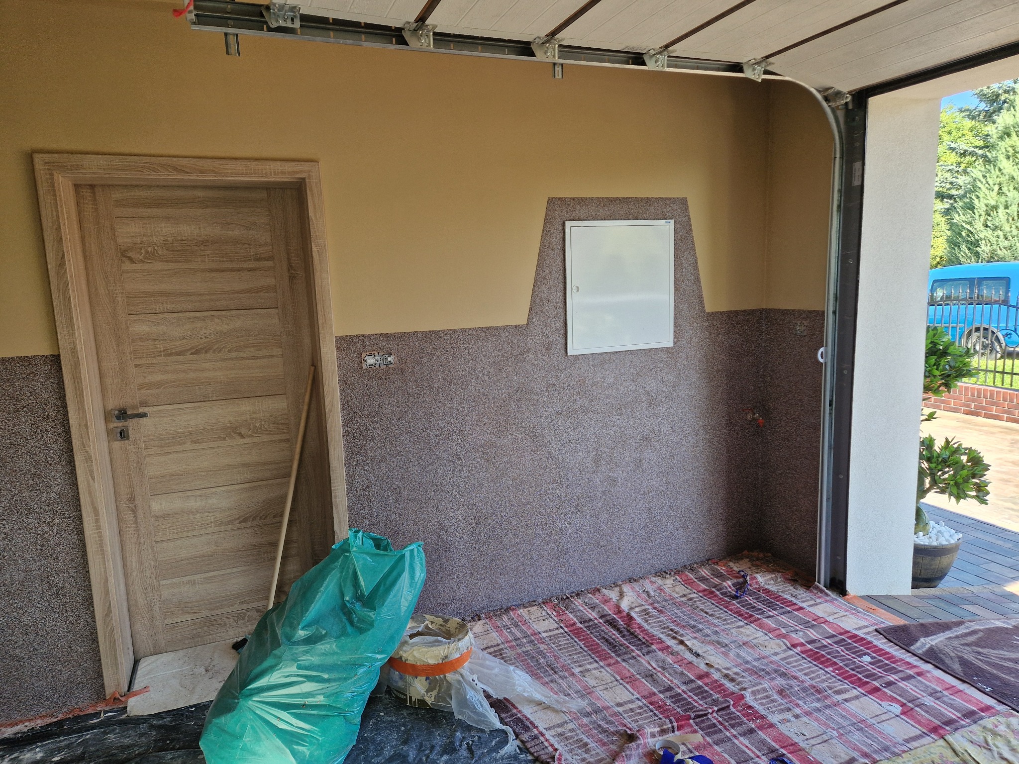 Wnętrze garażu z częściowo wykończonymi ścianami (struktura baranka) i nową, drewnianą ościeżnicą. Widoczny worek z odpadami i zabezpieczenie podłogi.