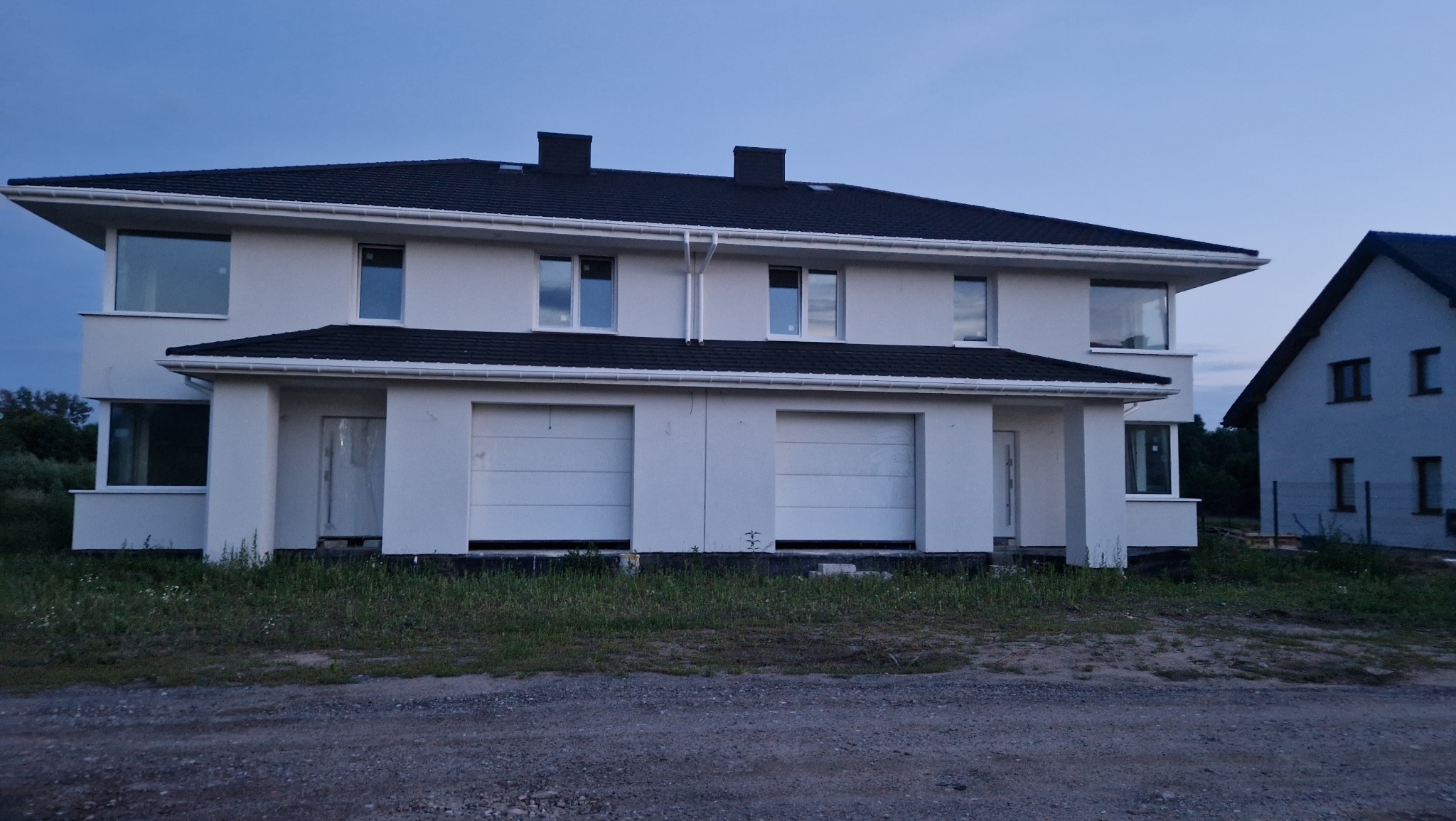 Dwukondygnacyjny, biały dom bliźniak z czarnym dachem i dwoma garażami, widziany z zewnątrz, w trakcie wykończenia. Widoczne okna i drzwi wejściowe.