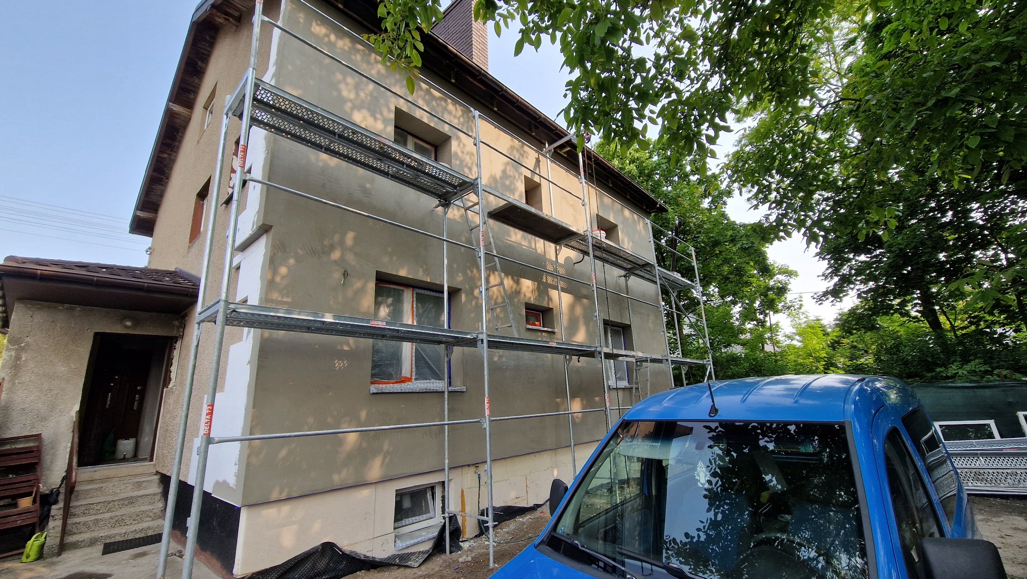 Dwupiętrowy dom w trakcie ocieplania, z rusztowaniem i częściowo otynkowaną elewacją. Widoczny fragment niebieskiego samochodu dostawczego i zieleń drzew.