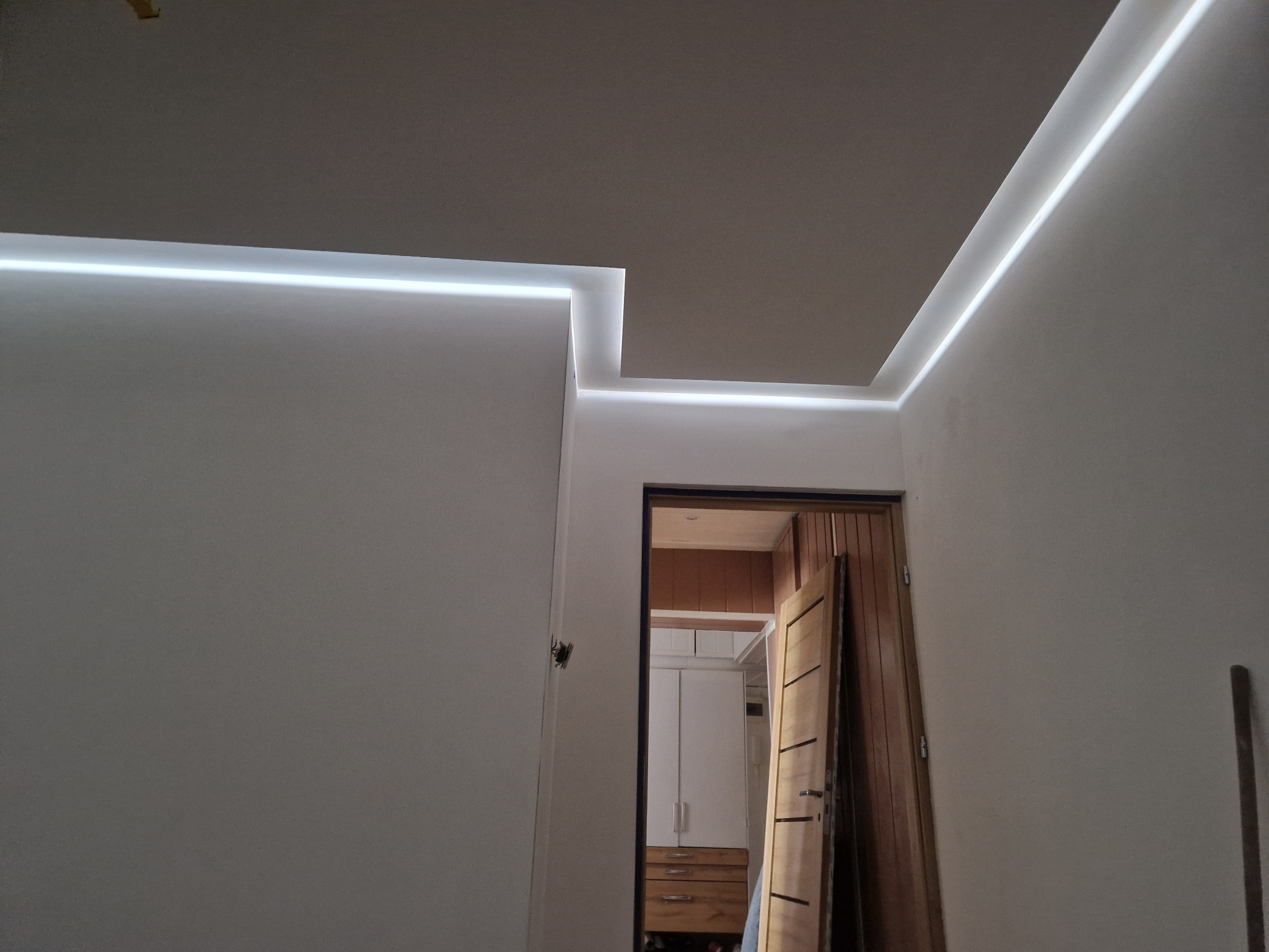 Nowoczesne oświetlenie LED wzdłuż krawędzi sufitu i ścian, tworzące minimalistyczny design. Widok na otwarte drzwi do pomieszczenia z szafą.