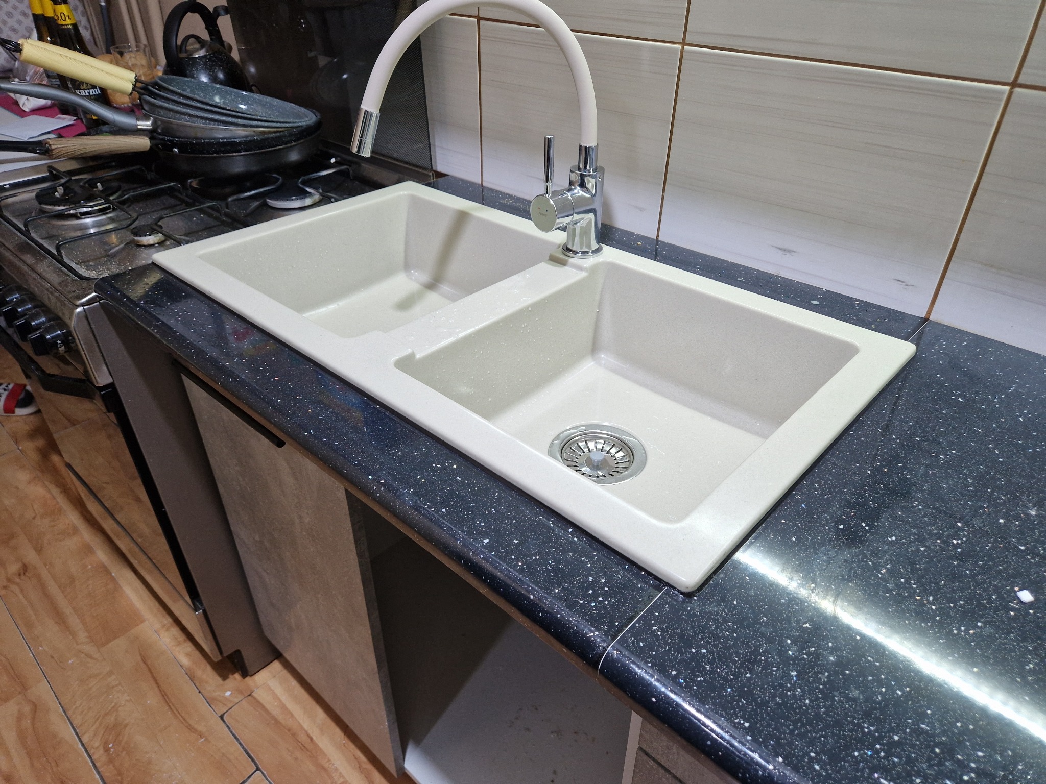 Dwukomorowy, beżowy zlewozmywak z baterią o białym, elastycznym wężu, osadzony w czarnym blacie z drobinkami, na tle płytek. Widoczny fragment kuchenki i szafki.