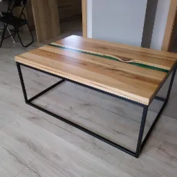 Stolik kawowy z blatem z jasnego drewna z żywicą epoksydową w kolorze zielonym, osadzony na czarnych, metalowych nogach o geometrycznym kształcie.