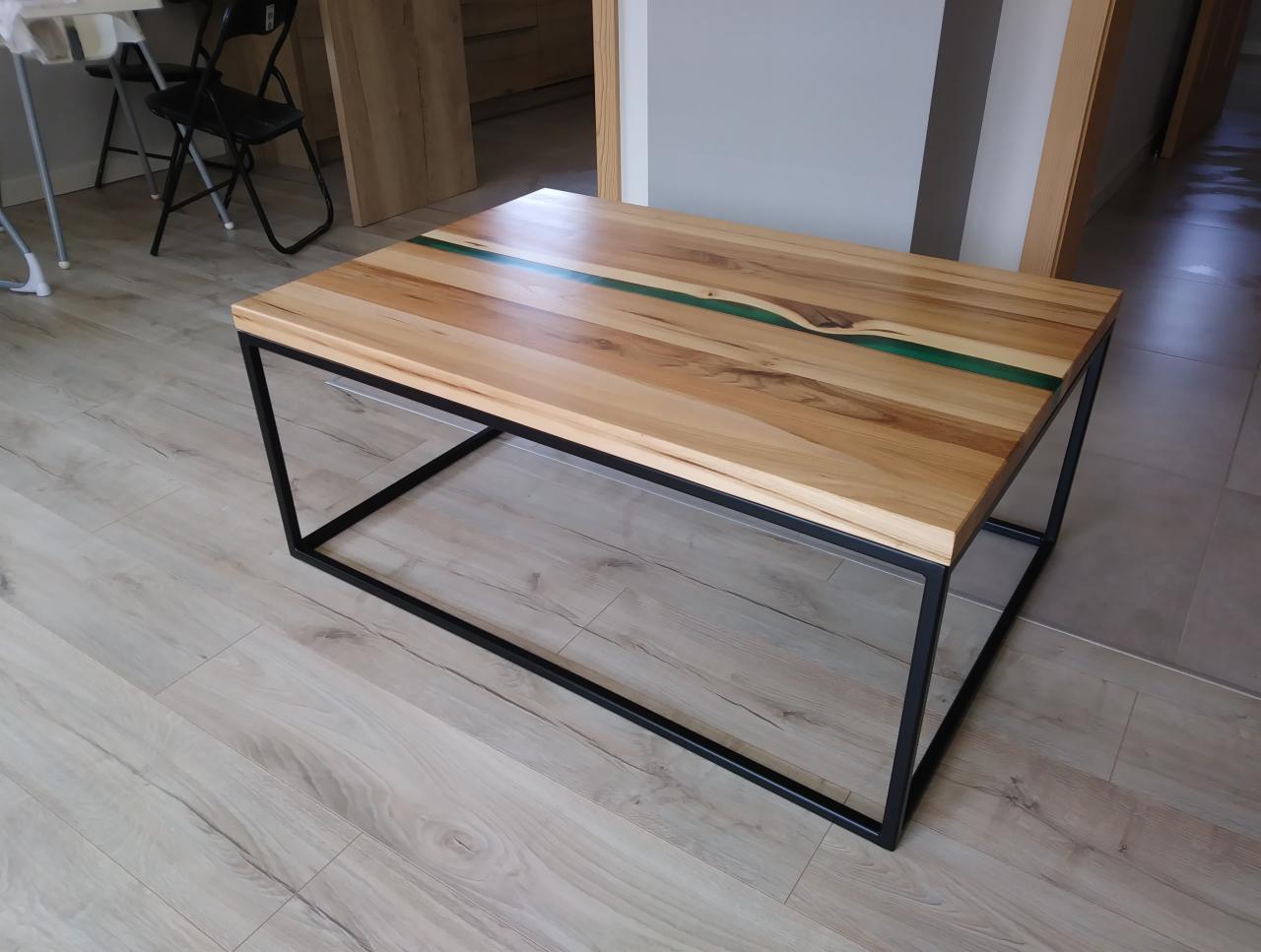 Stolik kawowy z blatem z jasnego drewna z żywicą epoksydową w kolorze zielonym, osadzony na czarnych, metalowych nogach o geometrycznym kształcie.