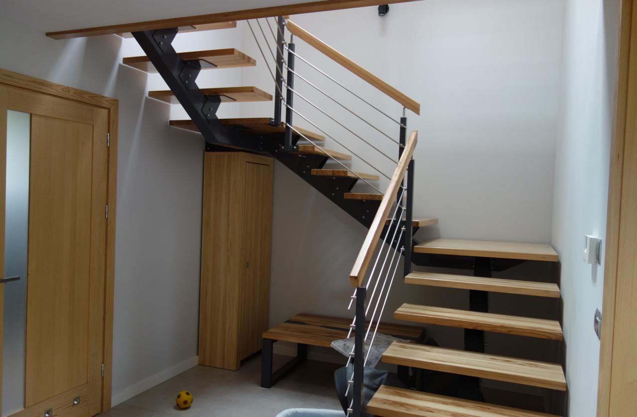 Nowoczesne schody z drewnianymi stopniami, metalową konstrukcją i balustradą ze stalowych linek, oświetlone od góry, z szafką pod biegiem schodów i dziecięcą piłką na podłodze.