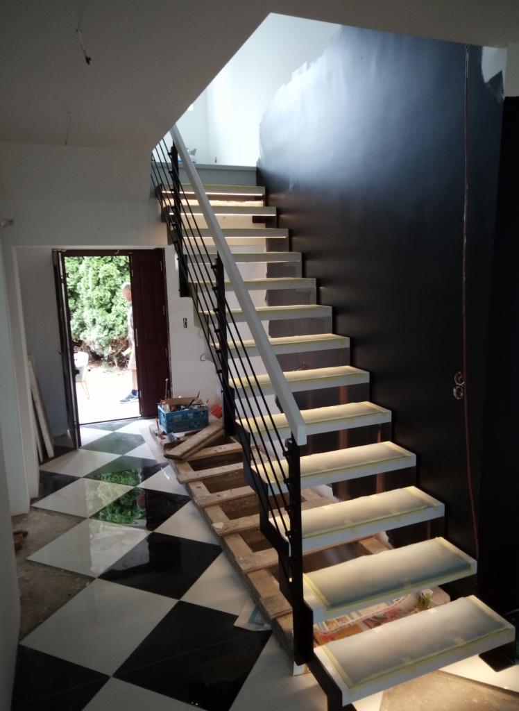 Montaż nowoczesnych schodów z podświetlanymi stopniami i czarną balustradą, widoczne zabezpieczenia na stopniach oraz szachownicowa podłoga w trakcie prac wykończeniowych.