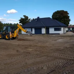 Żółta koparko-ładowarka JCB wykonuje prace ziemne na placu budowy nowego domu jednorodzinnego z ciemną dachówką, widoczne ślady opon na ubitej ziemi.