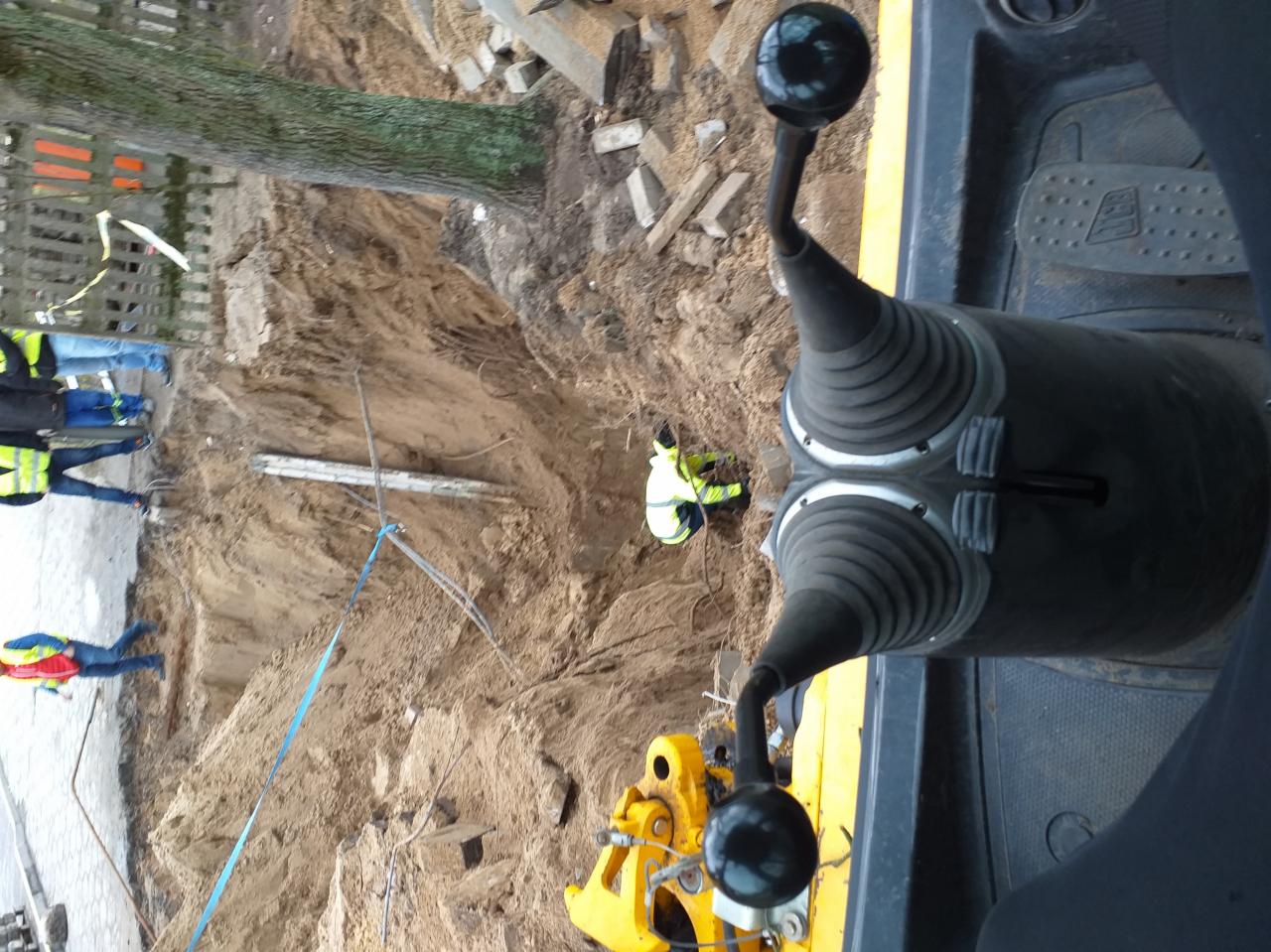 Widok z kabiny żółtej koparki na głęboki wykop w ziemi, gdzie pracuje osoba w odblaskowej kamizelce, z widocznymi fragmentami drzewa i innych osób w tle.