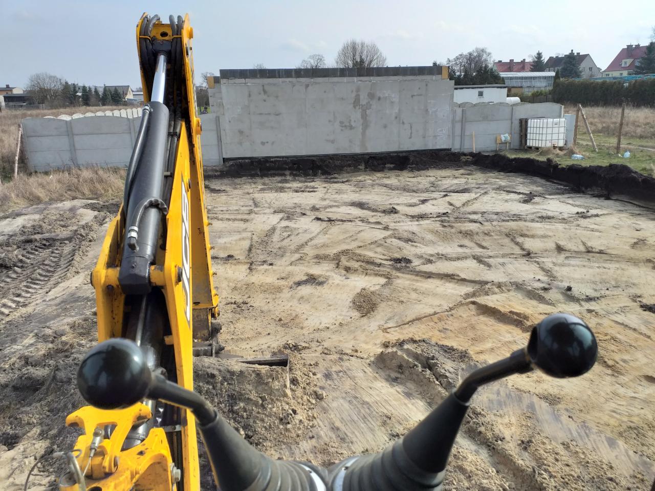 Widok z kabiny żółtej koparki JCB na przygotowany teren pod fundamenty, widoczny wykop z betonowym ogrodzeniem w tle.