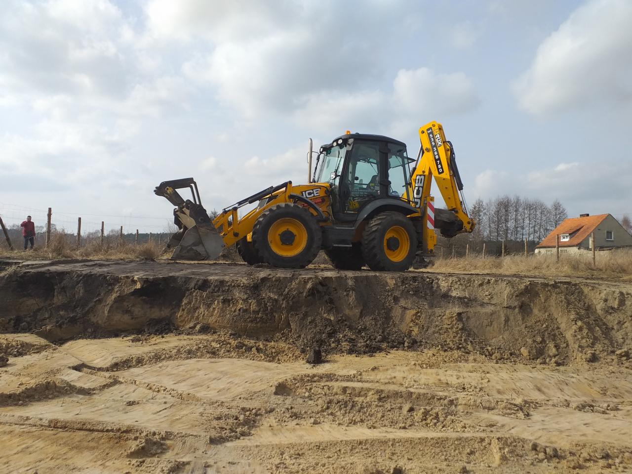 Żółta koparko-ładowarka JCB na skraju wykopu z widocznymi śladami pracy łyżki, w tle budynek mieszkalny i osoba obserwująca.