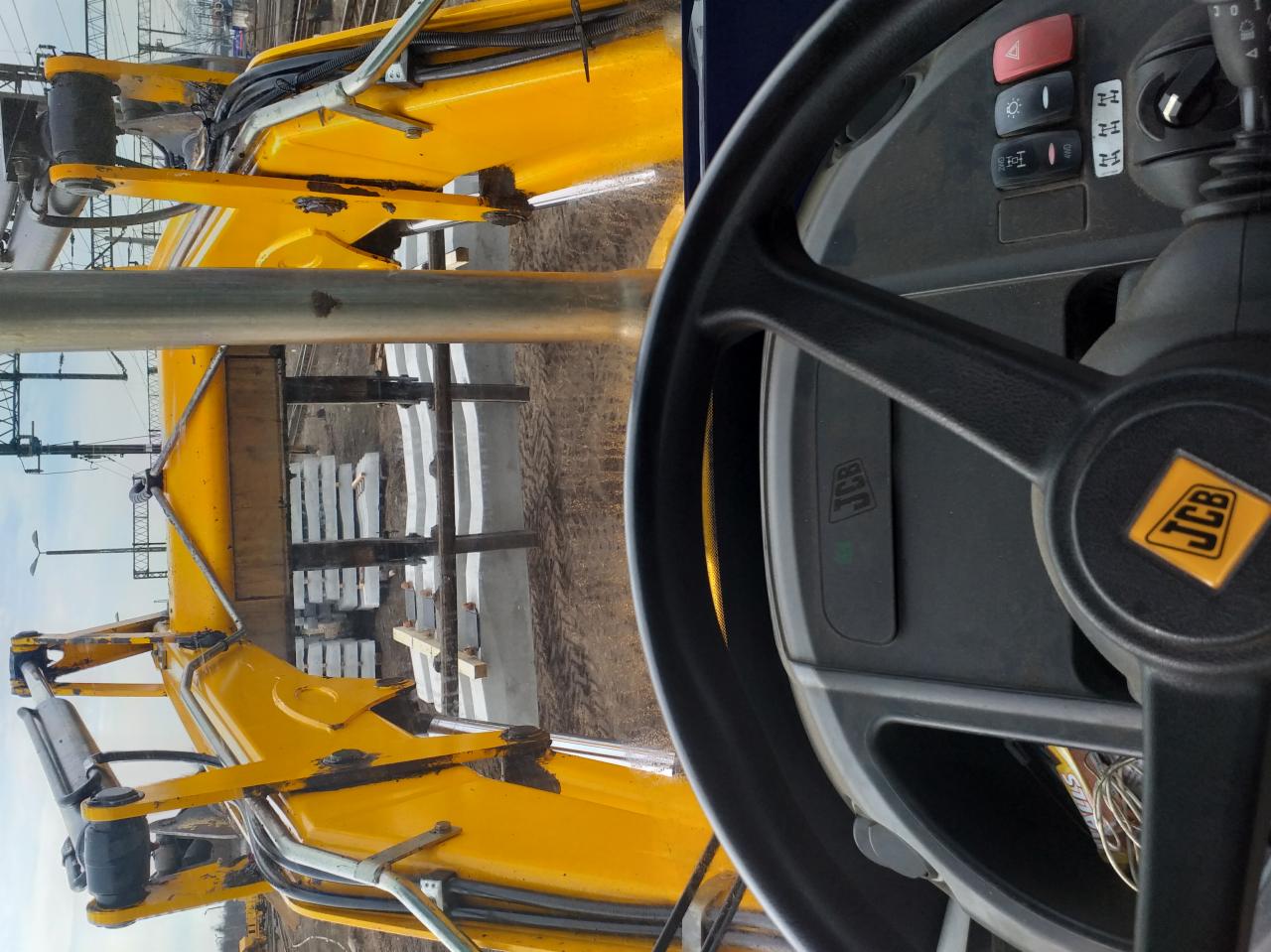 Widok z kabiny żółtej koparko-ładowarki JCB na plac budowy z przygotowanymi prefabrykatami betonowymi, w tle widoczne tory kolejowe.