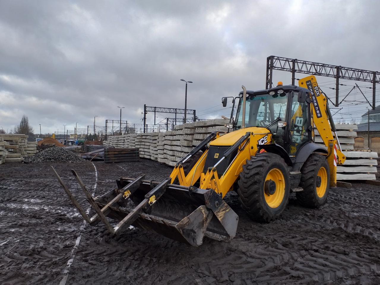 Żółto-czarna koparko-ładowarka JCB z widłami i łyżką, stojąca na błotnistym terenie budowy, w tle stosy betonowych elementów i konstrukcja stalowa